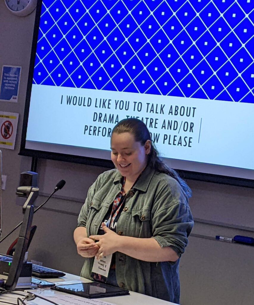 Photo of Sarah delivering a presentation. The slide behind her reads "I would like you to talk about drama, theatre, and/or performance now please".